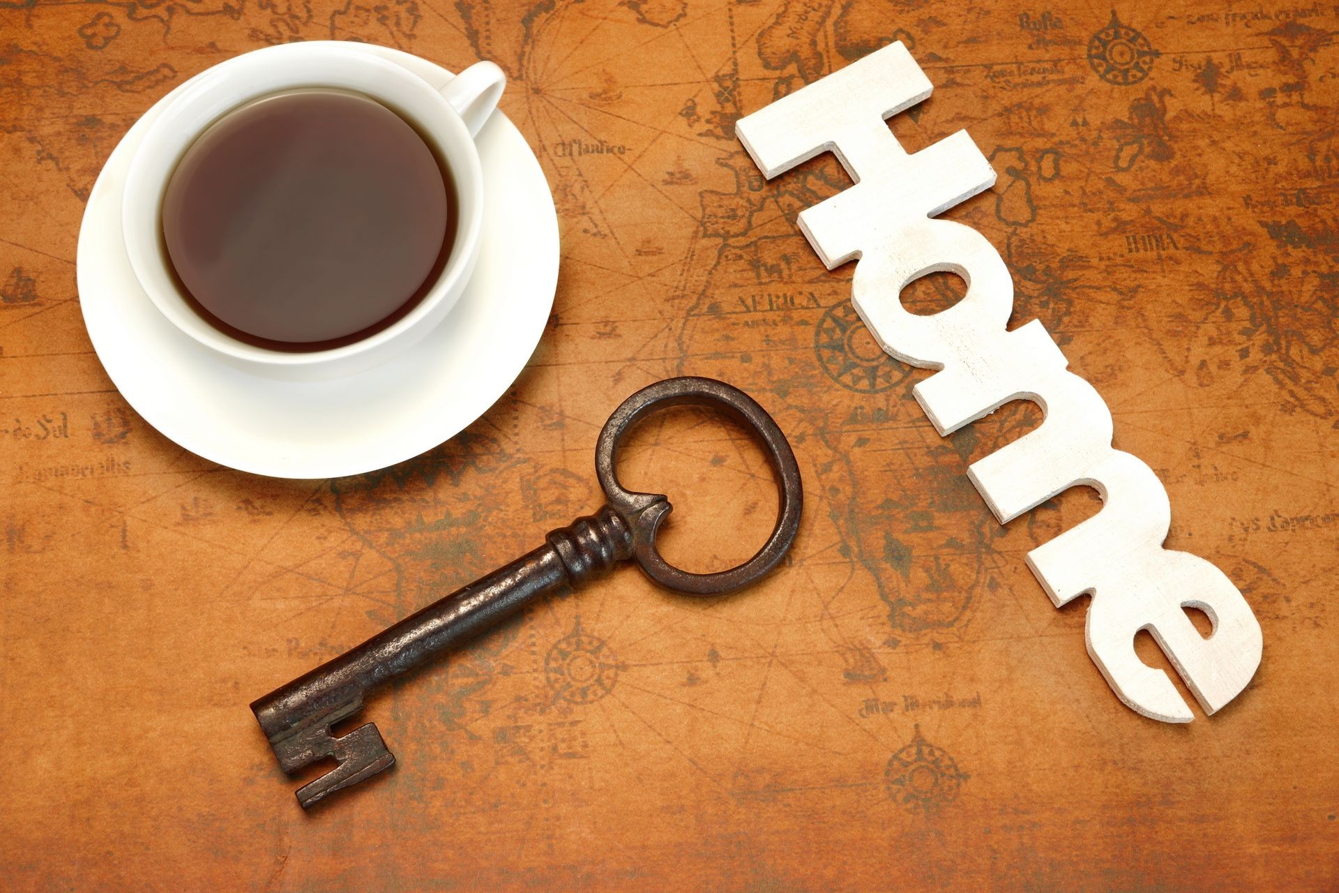 Dream Of Own Home Concept. White Tea Cup, Wooden Sign HOME And Retro Key On The Old World Map Background. Overhead View.